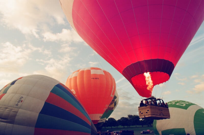 Several colorful hot air balloons, one with a bright flame, prepare for lift-off on a grassy field under a partly cloudy sky. People are visible in the nearest wooden basket.