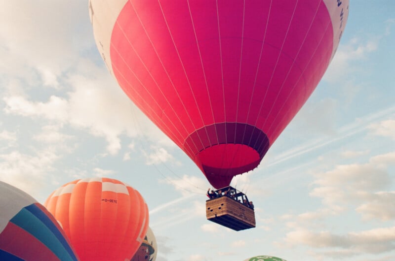 A large red hot air balloon with people in the basket floats in the sky, surrounded by other colorful balloons, against a backdrop of clouds.