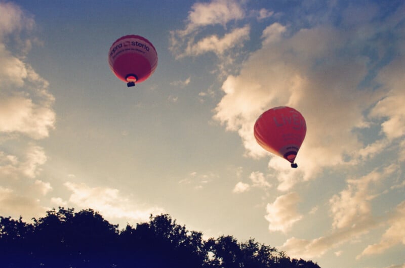 Two hot air balloons float in a partly cloudy sky above tree silhouettes at sunset or sunrise.
