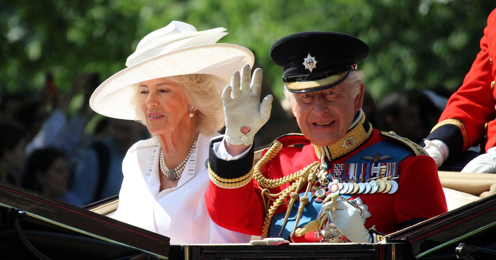 A man in a decorated military uniform waves from a carriage beside a woman in a white hat and outfit, both participating in a formal outdoor event with a blurred crowd in the background.