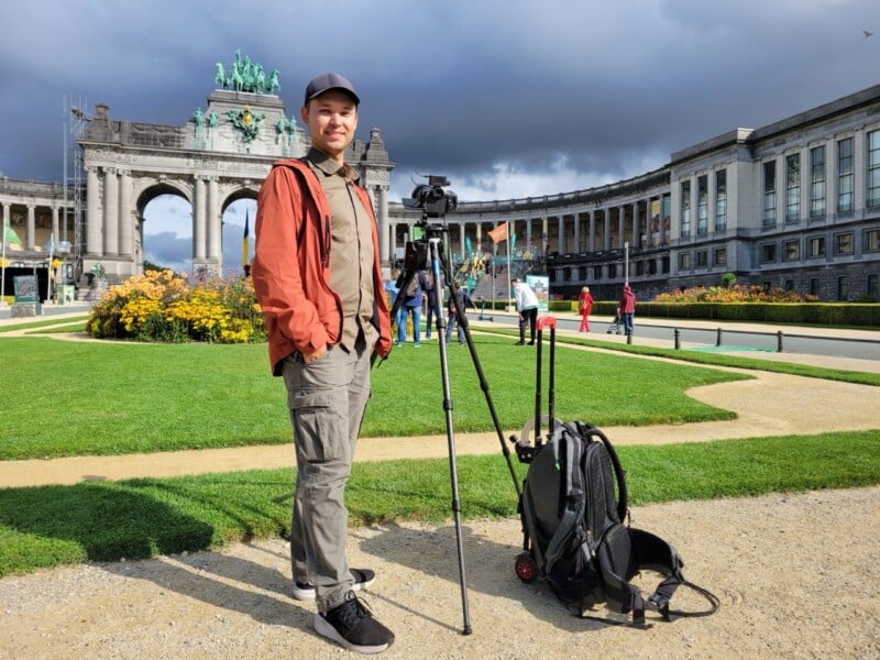 A person stands next to a camera on a tripod in a park with an arch monument and curved building in the background, under a cloudy sky. A backpack and another tripod are on the ground nearby.