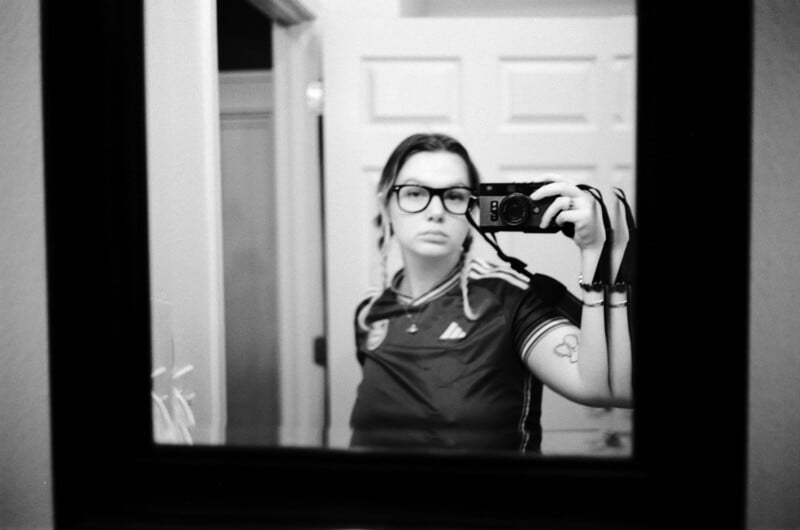 A person with braided hair and glasses takes a mirror selfie while holding a camera, wearing a short-sleeved athletic shirt.