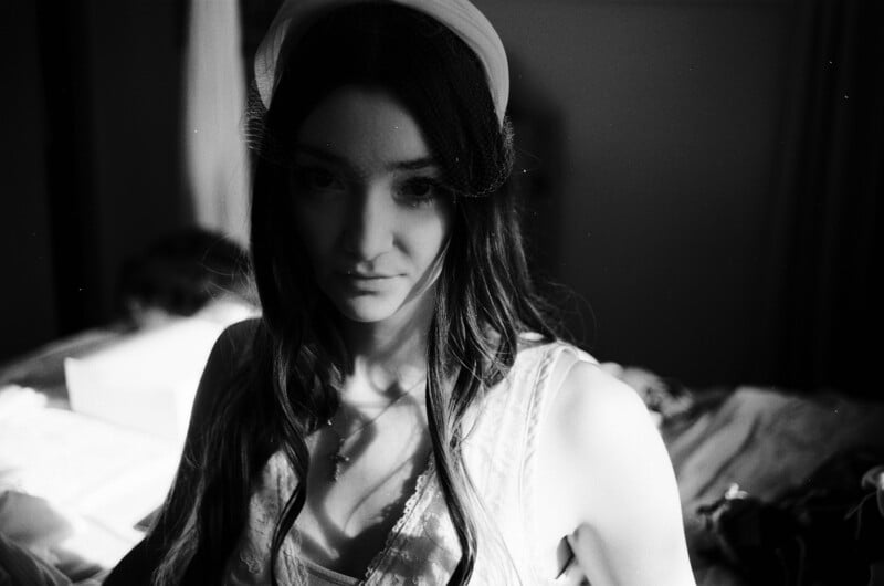 A young woman with long dark hair wears a white headband with a veil, looking directly at the camera in a dimly lit room.