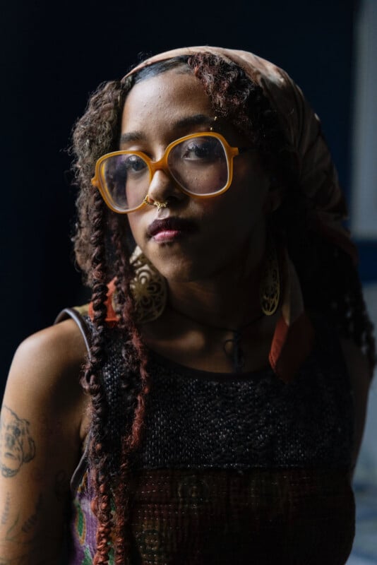 A young woman with curly hair, wearing large yellow glasses, a headscarf, large earrings, and a nose ring, looks at the camera. Tattoos are visible on her arm. The background is dark and blurred.