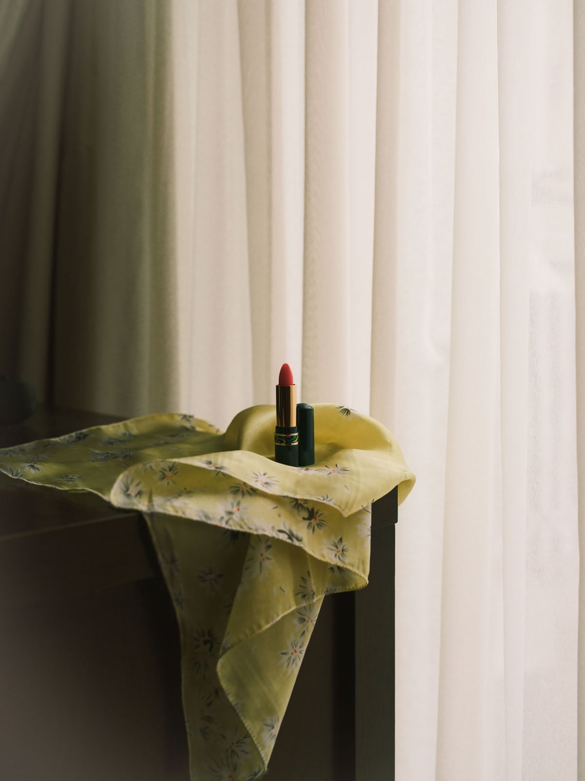 A red lipstick stands upright on a yellow floral-patterned cloth draped over a dark surface in front of white curtains.