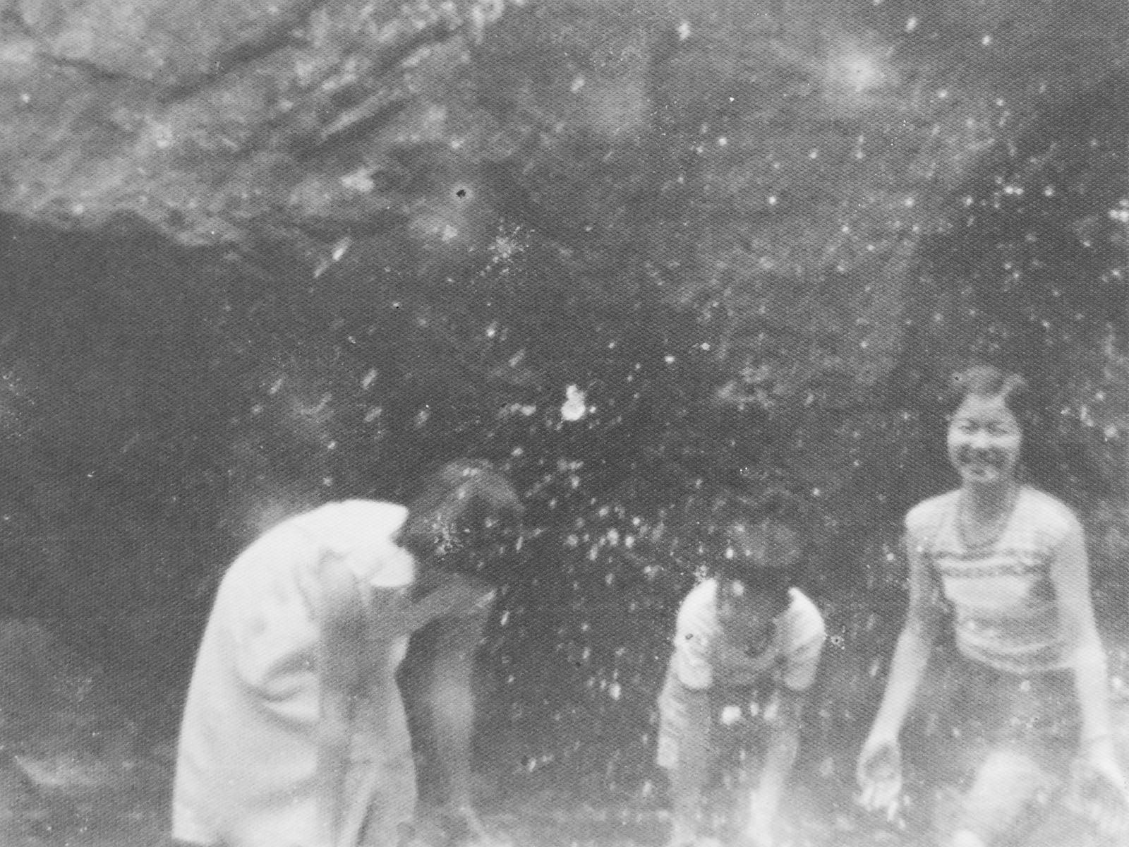 Three people stand in shallow water, with one person on the left leaning over and two others standing upright.