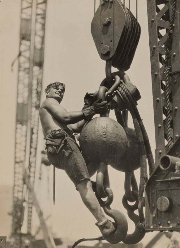 A shirtless construction worker wearing gloves and shorts hangs from the heavy metal hook and ball assembly of a large crane.