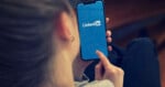 A person holds a smartphone displaying the LinkedIn logo on its screen while touching the display with their index finger.