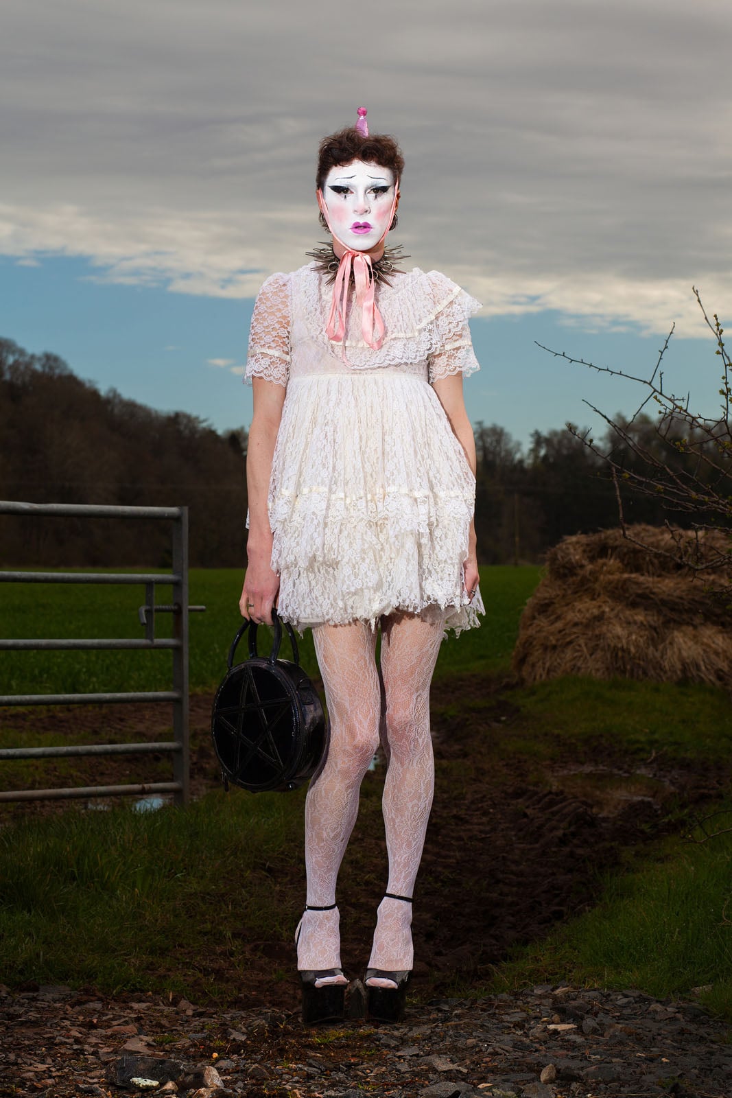 A person in a white lace dress, lace tights, platform shoes, dramatic makeup, and a pink bow stands outdoors on a dirt path, holding a black handbag. The sky is cloudy and there's grass, a metal gate, and hay in the background.