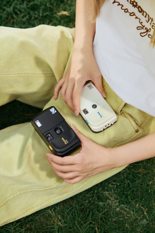 Person sitting on grass holds two Kodak film cameras, one black and one white, resting on their yellow pants. Only their hands and torso are visible; they wear a white shirt with cursive writing.