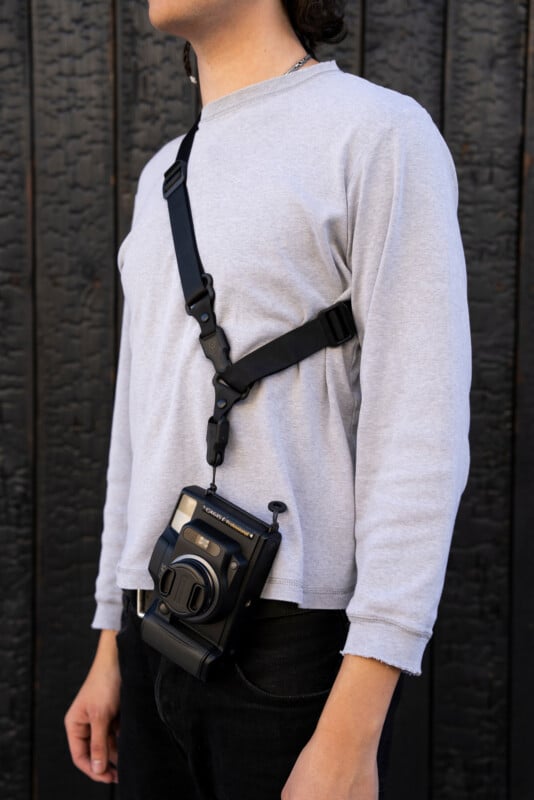 Person wearing a light gray long-sleeve shirt and black pants, standing against a dark textured wall, with a black camera strapped across their chest. The person's face is partially out of frame.