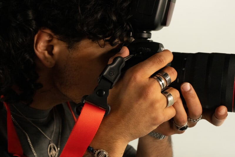 A person with curly hair and multiple rings holds a camera up to their eye, ready to take a photo. The camera has a red strap, and the background is neutral.