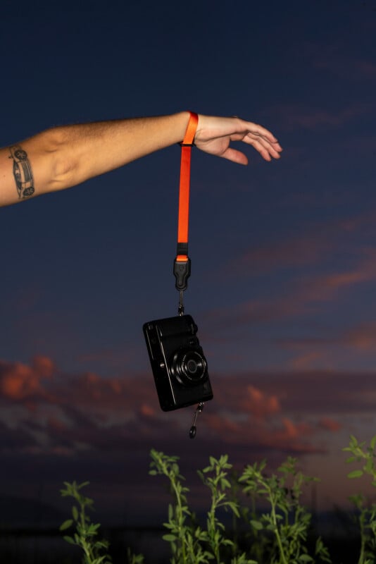 An outstretched arm with a tattoo holds a black camera by an orange wrist strap over green plants at dusk, with a dark blue and pink sky in the background.