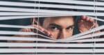 A person peers through open blinds with both hands, looking intently outside.