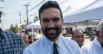 A man with dark hair and a beard, smiling, stands outdoors at an event with people and tents in the background.
