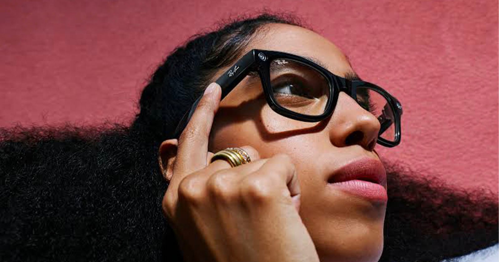 A person with long curly hair and glasses looks upward thoughtfully, touching the side of their black-framed glasses. Their lips are slightly parted and the background is a solid pink color.