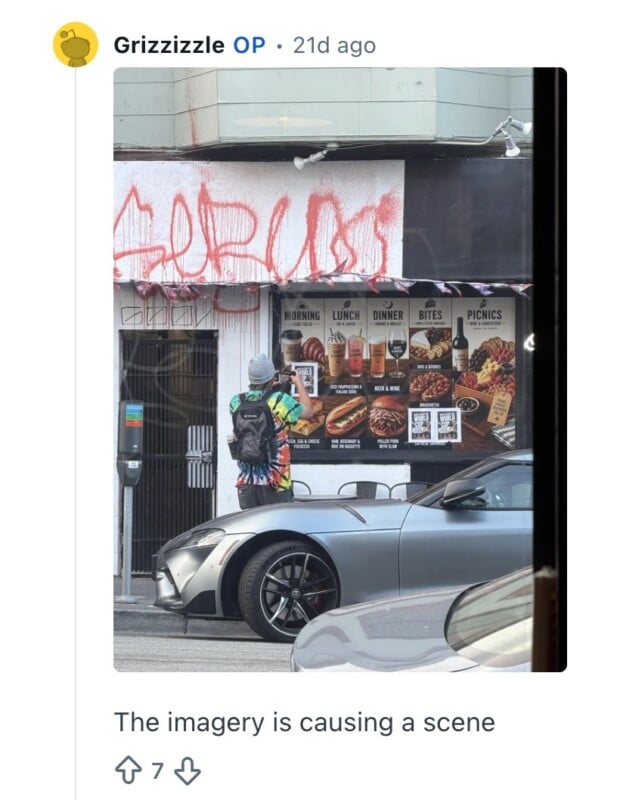 A person in colorful clothes and a yellow backpack takes a photo of a wall covered in red graffiti above food posters, with a silver car parked in front.
