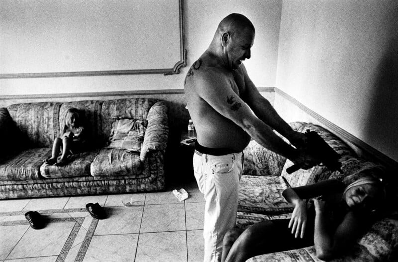 A shirtless man points a hairdryer at a reclining woman on a couch, while a child sits alone on another couch in a dimly lit living room.