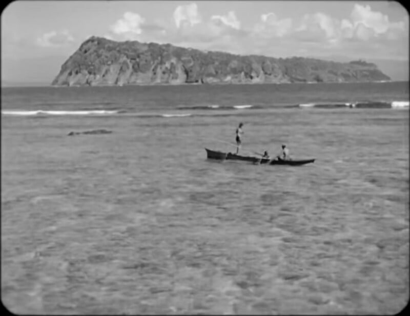 Three people are in a small wooden boat on calm, shallow water with an island and scattered clouds visible in the background.