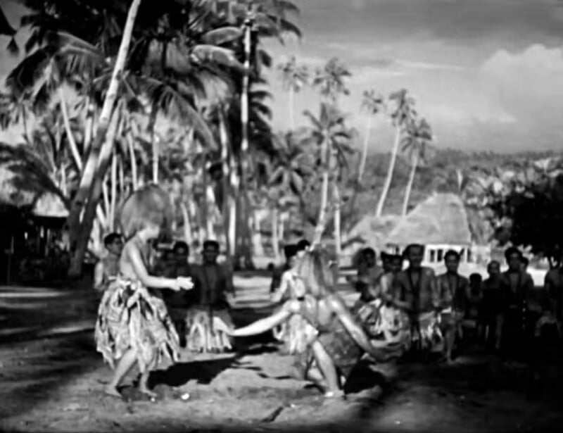 Two people in traditional costumes dance on a sandy area, surrounded by a group watching. Palm trees and huts are visible in the background. The image is in black and white.