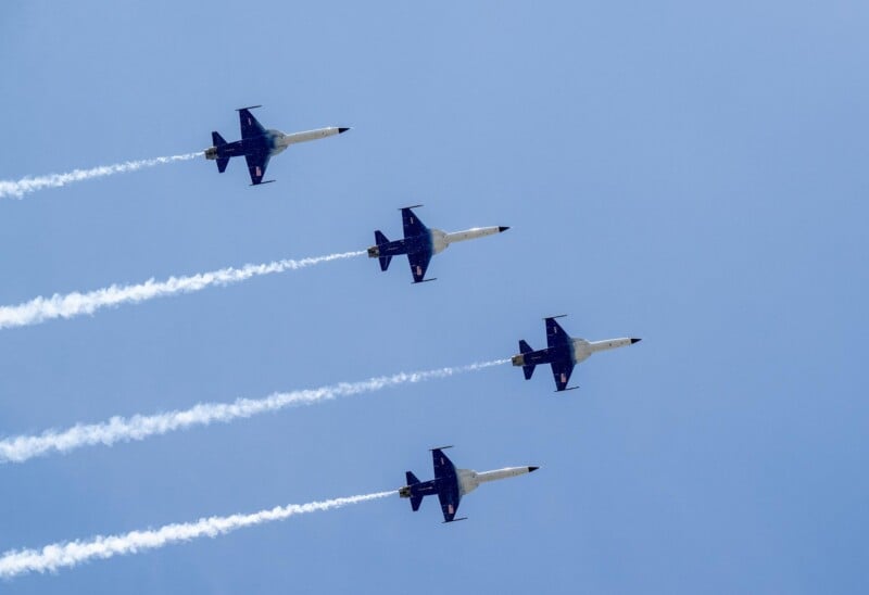 Four fighter jets fly in a tight formation against a clear blue sky, leaving white vapor trails behind them.