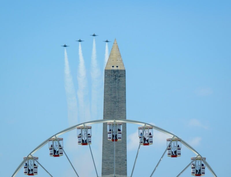 Four jets fly in formation leaving white trails behind the Washington Monument, with the upper portion of a Ferris wheel visible in the foreground against a clear blue sky.