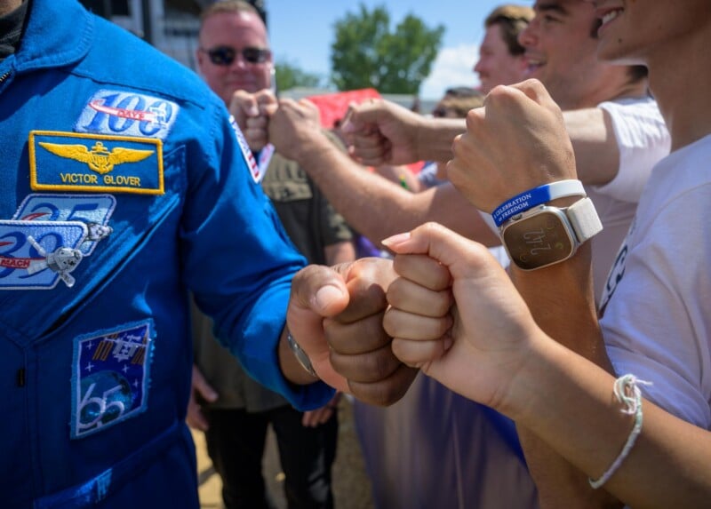 A group of people fist bumps an astronaut in a blue suit with the name “Victor Glover” visible on a patch. Others wear casual clothes, bracelets, and a smartwatch. The scene is outdoors on a sunny day.