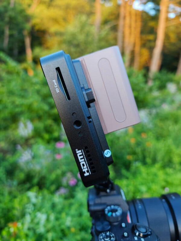 Close-up of a digital camera with an external monitor attached, displaying an SD card slot. The background shows a green, blurry outdoor setting with trees and foliage.