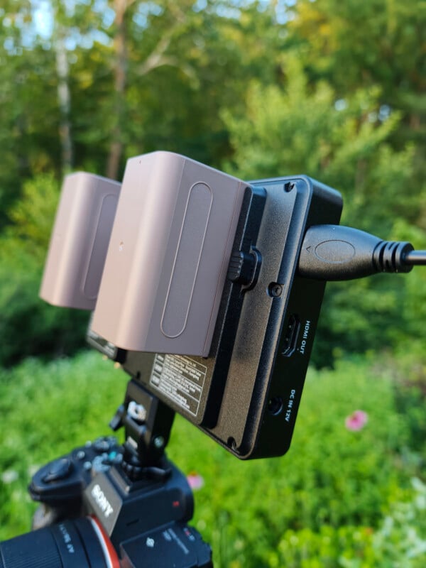 Close-up of a camera with two large batteries attached and a cable plugged in, set outdoors with greenery and blurred flowers in the background.