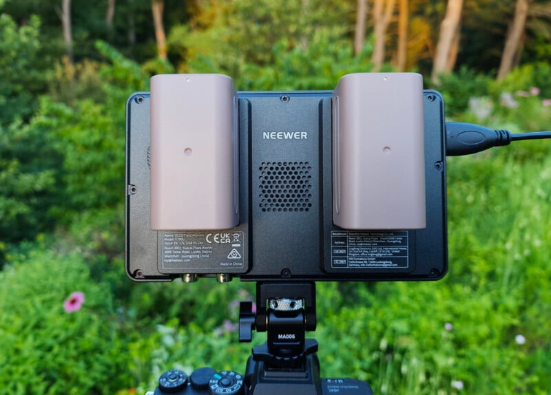A Neewer device with two large batteries attached is mounted on a tripod outdoors, with a green, blurry garden and trees in the background.