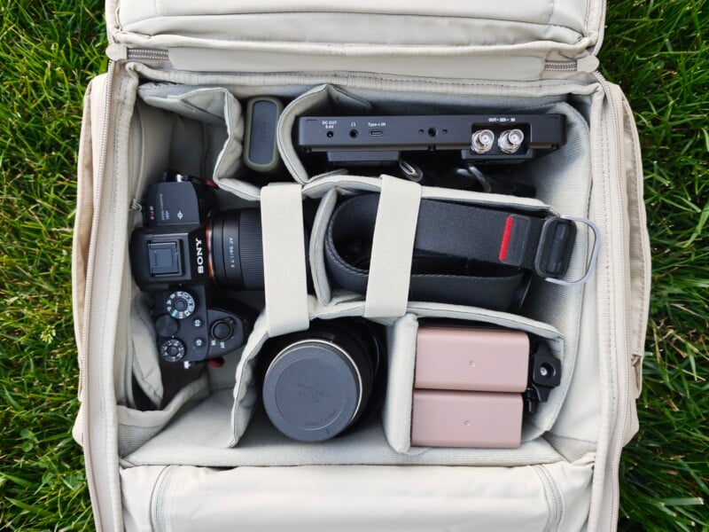 Open camera bag on grass containing a Sony camera, two lenses, two batteries, camera strap, and a rectangular electronic device organized in padded compartments.