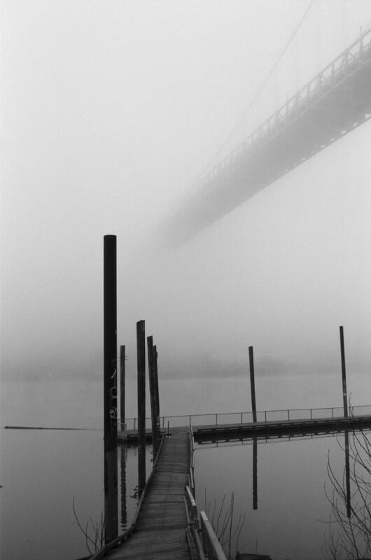 A foggy scene shows a wooden dock extending over calm water, with tall posts rising alongside. A large bridge fades into the mist above, creating a mysterious and quiet atmosphere.