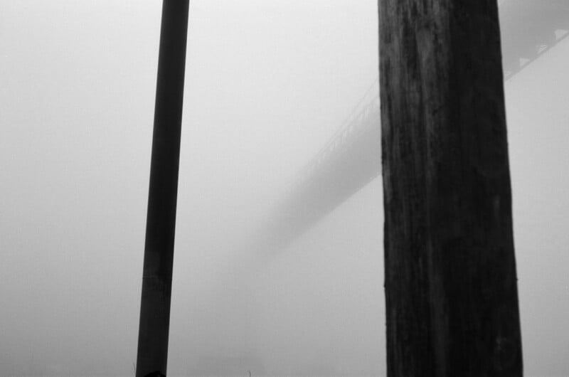 Two vertical poles stand in the foreground, partially obscuring a bridge that fades into heavy fog, creating a mysterious and atmospheric scene with limited visibility.