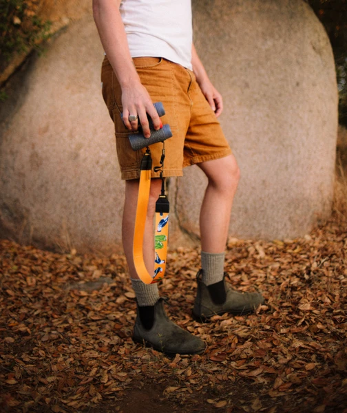 A person in brown shorts and boots stands on dry leaves, holding binoculars and a water bottle with a yellow strap. Large rocks are in the background.