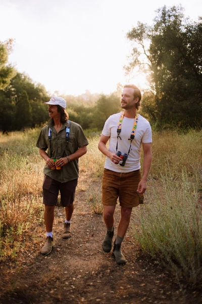 Two people walk on a dirt trail through a grassy, sunlit field, each holding binoculars. They are casually dressed and appear to be enjoying a nature walk surrounded by trees and tall grass.