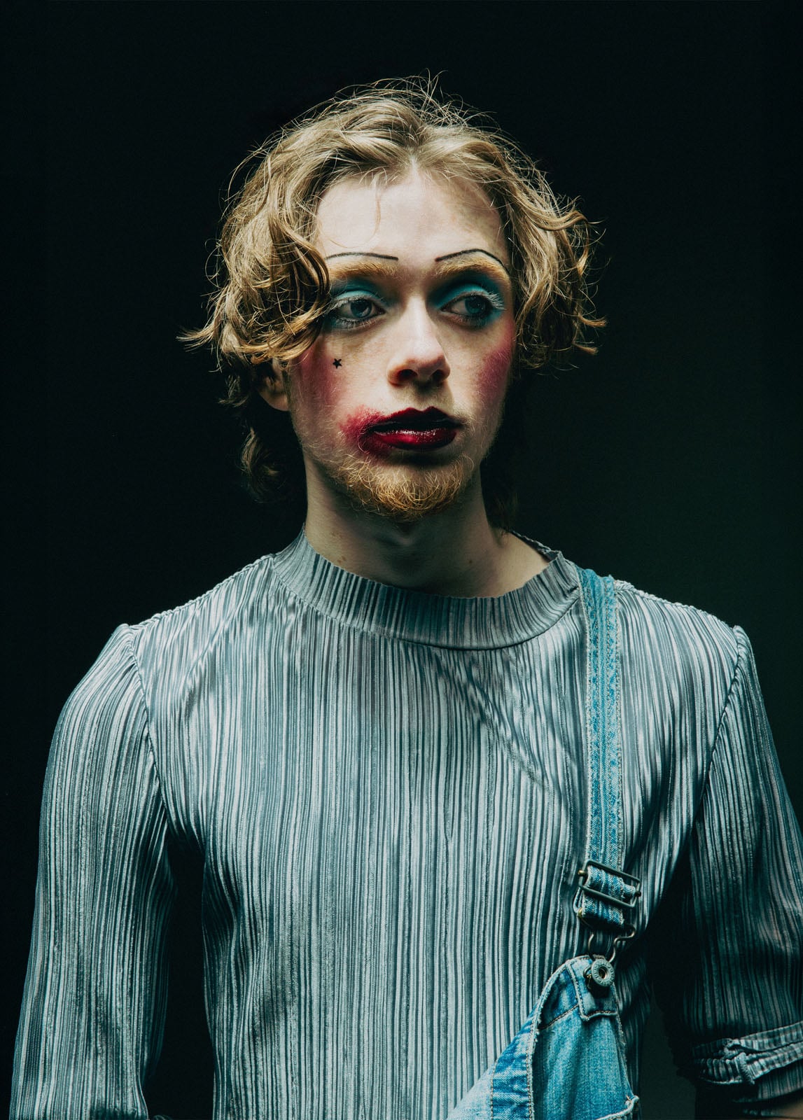 A person with curly hair wears dramatic makeup, including bold eyeshadow, dark lipstick, and a beauty mark. They have a short beard and mustache, and wear a pleated top with one strap of denim overalls.