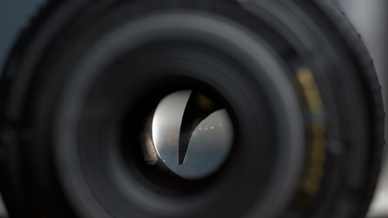 Close-up view of a camera lens aperture, showing the curved blades partially closed with reflections of light visible on the inner glass elements.