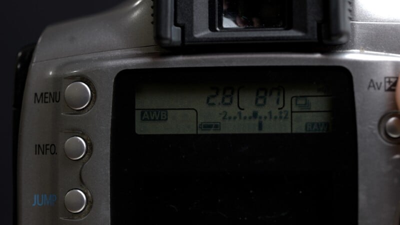 Close-up of a digital camera's control panel and LCD screen displaying camera settings. The screen shows “2.8 (87),” along with icons for AWB, battery level, and RAW format. Menu, info, and jump buttons are visible on the left.