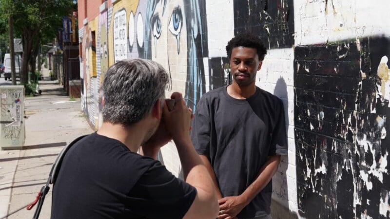 A man poses against a graffiti-covered wall while another man in front of him takes his photo on a sunny city sidewalk.