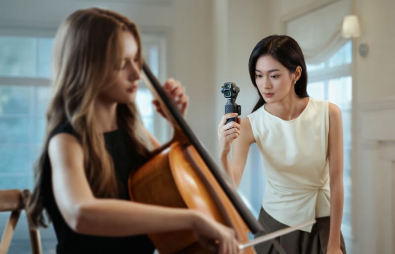 A woman in a white top holds a small, handheld gimbal camera while filming a woman playing a cello.