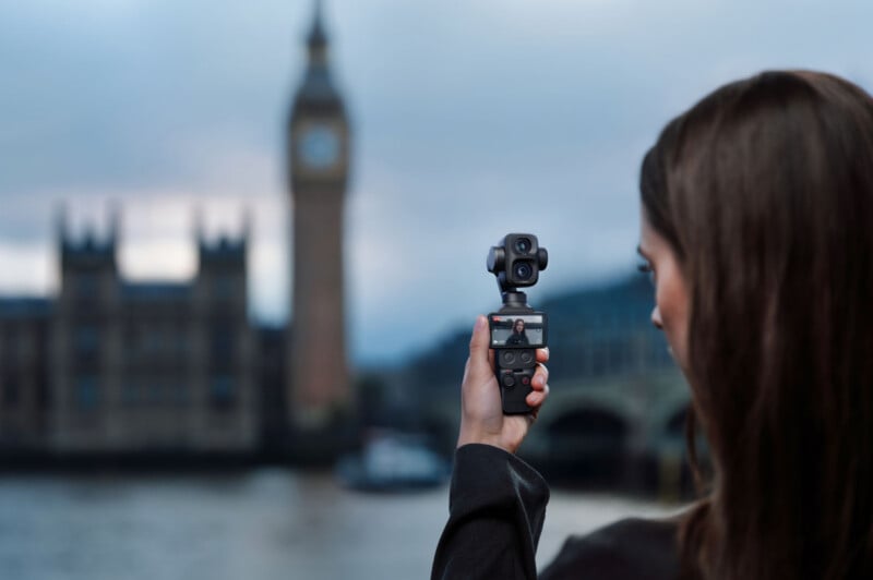 A person holds a small handheld gimbal camera, filming themselves with the blurred silhouette of Big Ben and the Houses of Parliament in the background.