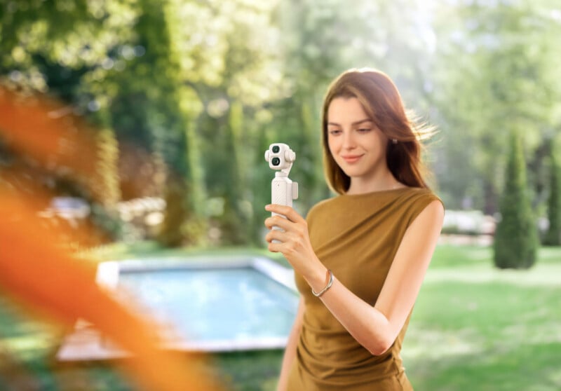 A woman in a brown dress holds a small, white handheld gimbal camera in a sunny outdoor setting with a swimming pool in the background.