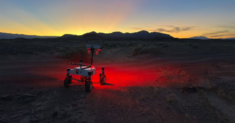 A small rover with lights glowing red underneath stands on rugged terrain at dusk, with mountains silhouetted against a colorful sunset sky in the background.