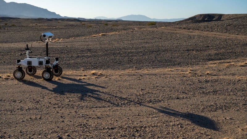 A small white rover with cameras and sensors stands on rocky desert terrain, casting a long shadow in the sunlight. Distant mountains are visible under a clear sky.