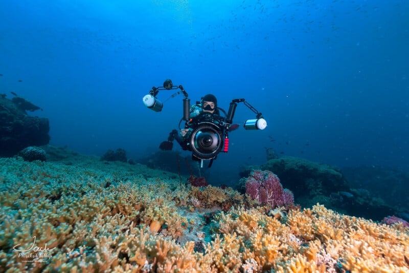 A scuba diver underwater holds a large camera with lights, photographing coral reefs and small fish swimming in the blue ocean background.