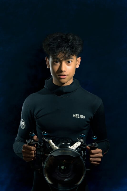 A young person with curly hair in a dark wetsuit holds a professional underwater camera against a dark blue background, looking directly at the camera.