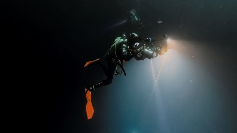 A scuba diver with orange fins, holding a camera and flashlight, swims in dark water with a beam of light illuminating the surroundings.