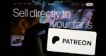 A hand holds a phone displaying the Patreon logo in front of a screen with the text 