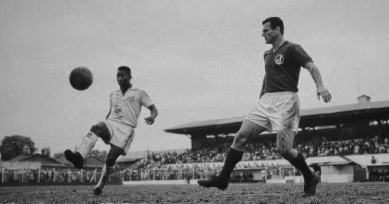 A black-and-white photo shows two soccer players on a field, one kicking the ball mid-air as the other approaches. Spectators fill the stands in the background.