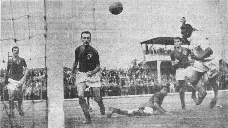 Black and white photo of a soccer match, with players in action near the goal, one on the ground, a ball in mid-air, and a crowd watching from the stands in the background.